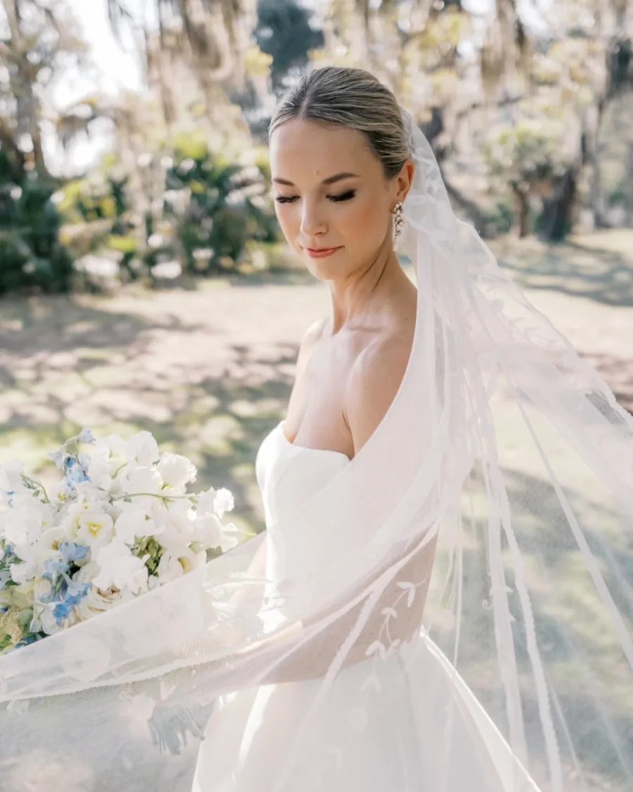 A bride in a veil holding a bouquet, beneath live oaks.